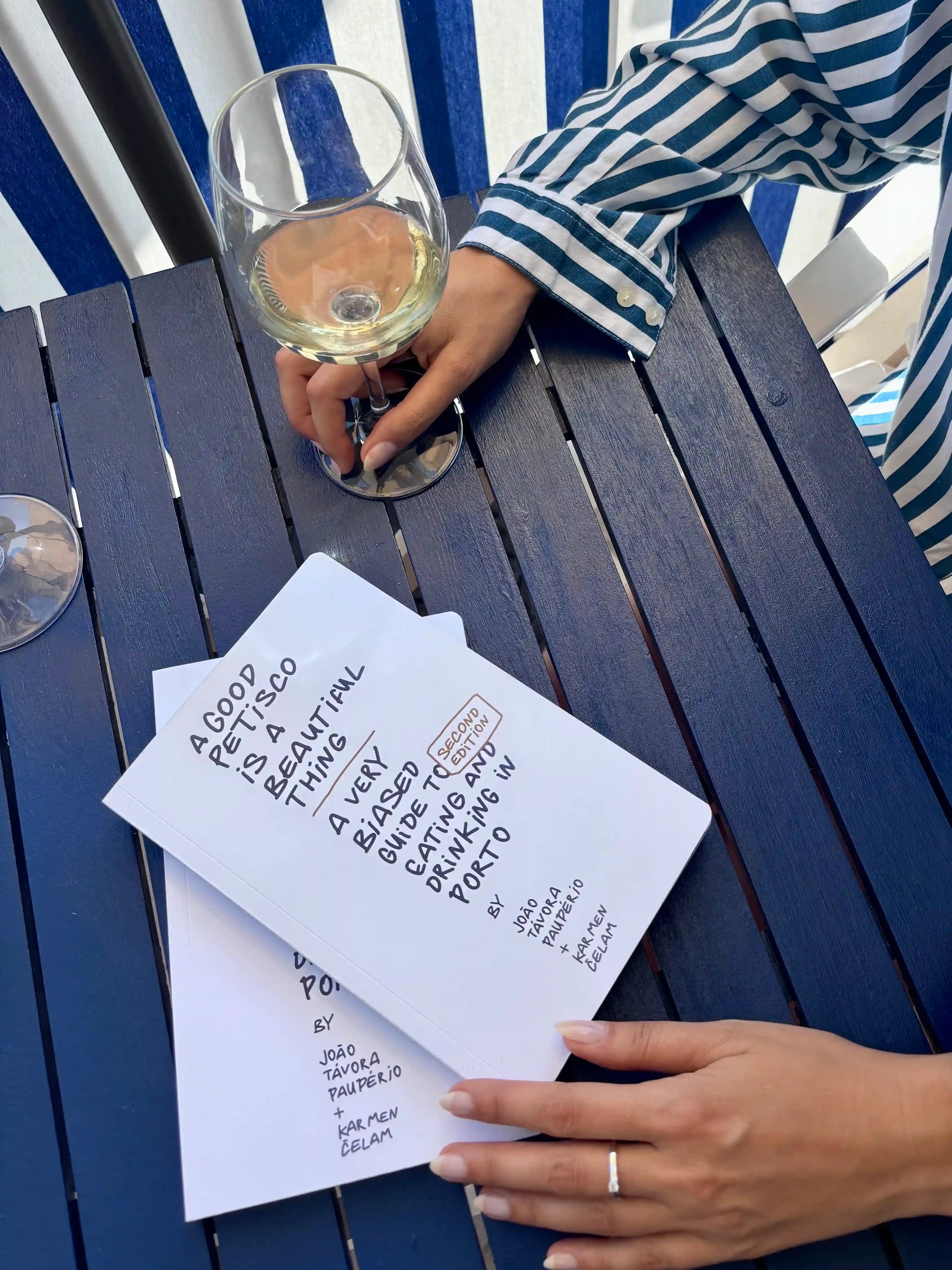 Person holding a glass of white wine with a menu on a blue table outdoors.