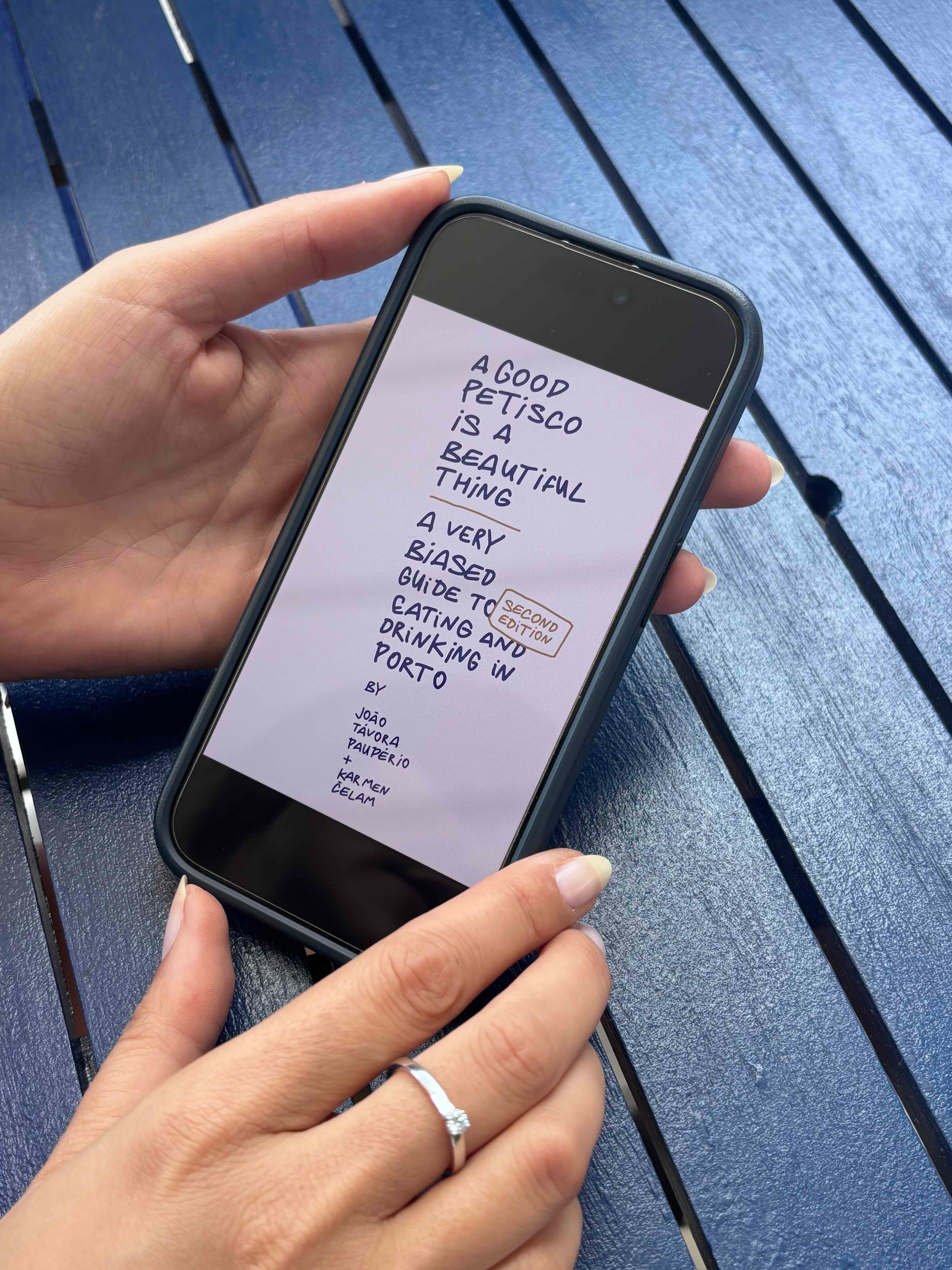 Person holding a phone with The Petiscos ebook on a blue wooden surface